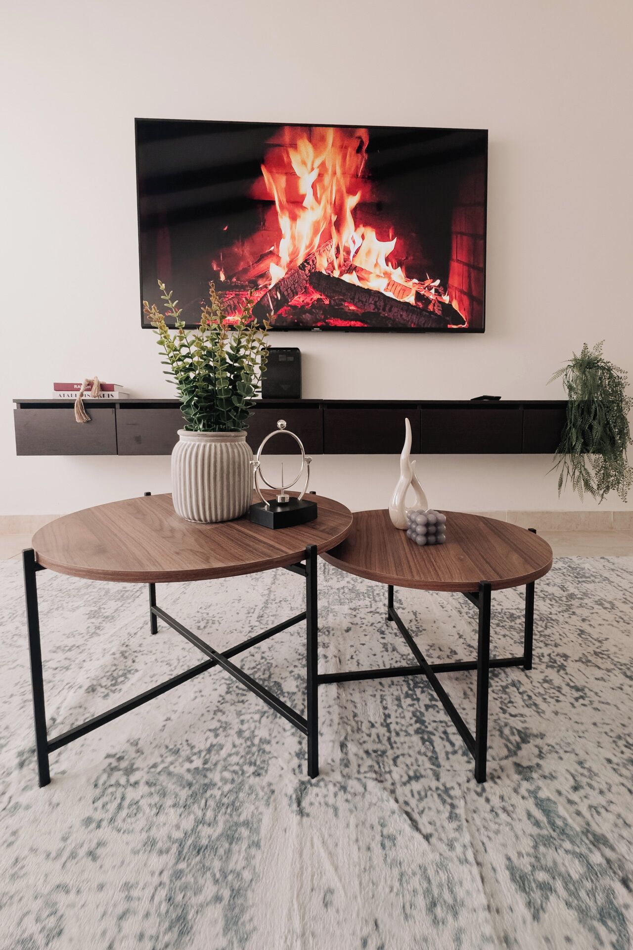 Een moderne woonkamer met een aan de muur bevestigde televisie, twee ronde houten tafels met metalen poten, een potplant en een decoratieve vaas.
