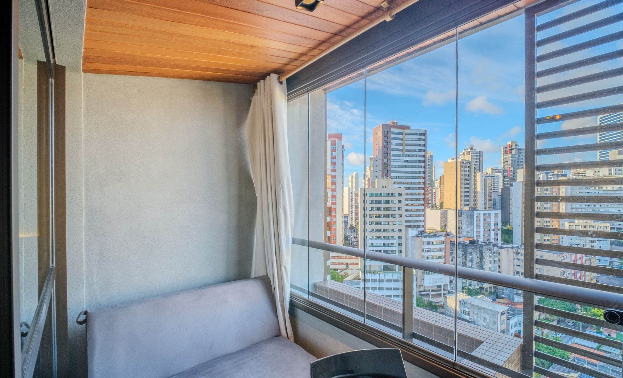 A balcony with a view of a cityscape, a cushioned seating area, and a wooden ceiling.