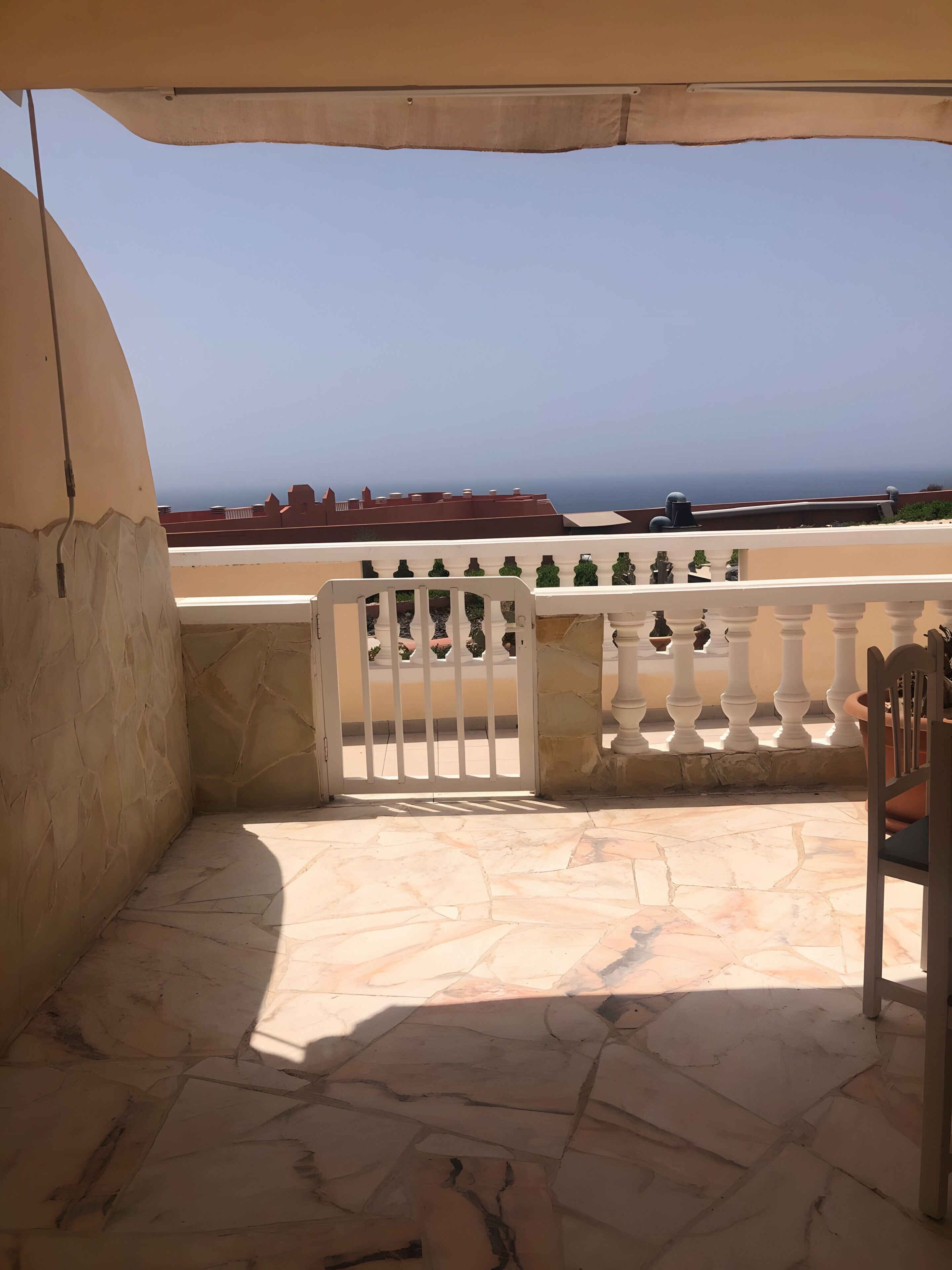 A terrace with a white balustrade, a stone floor, and a view of the sea and buildings in the distance.