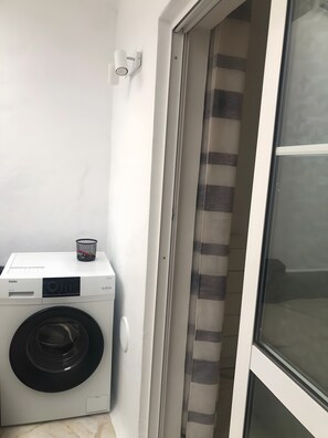 A compact laundry area with a front-loading washer and dryer, a small basket on the dryer, and a sliding glass door leading to an outdoor area. - Vacation Apartment 'Sotavento' with Sea View, Private Terrace and Wi-Fi (Pájara)