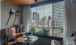 A room with a dining table set for two, a window with blinds, and a view of city buildings. - Studio 750m From the Sea, Building w/ Pool Eub1601 (Salvador)