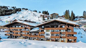 Un immeuble en bois à plusieurs étages, doté de balcons, situé sur une pente enneigée, avec des remontées mécaniques en arrière-plan.