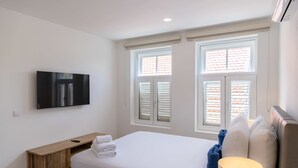 Hotellihuone, jossa on sänky, seinälle asennettu televisio, yöpöytä, jossa on kaukosäädin, ja kaksi ikkunaa, joissa on sälekaihtimet.