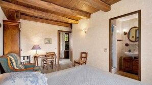 A bedroom with a wooden ceiling, a bed with a quilted cover, a chair, a table with a lamp, and a bathroom visible through an open door.