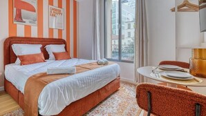 A hotel room with a bed, a dining table with chairs, a window with curtains, and a view of buildings outside.