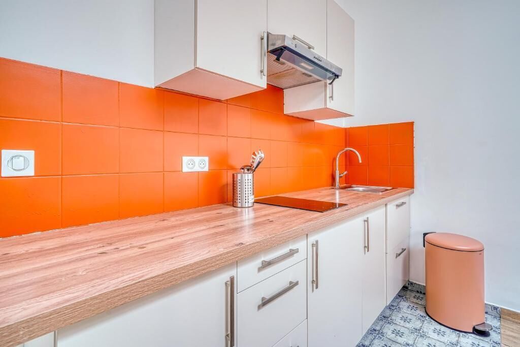 Een keuken met witte kastjes, een houten aanrechtblad, een oranje spatwand, een roestvrijstalen spoelbak en een prullenbak.