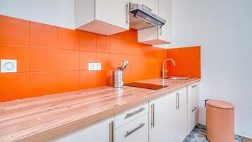 Een keuken met witte kastjes, een houten aanrechtblad, een oranje spatwand, een roestvrijstalen spoelbak en een prullenbak.