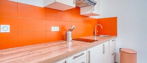 Een keuken met witte kastjes, een houten aanrechtblad, een oranje spatwand, een roestvrijstalen spoelbak en een prullenbak.
