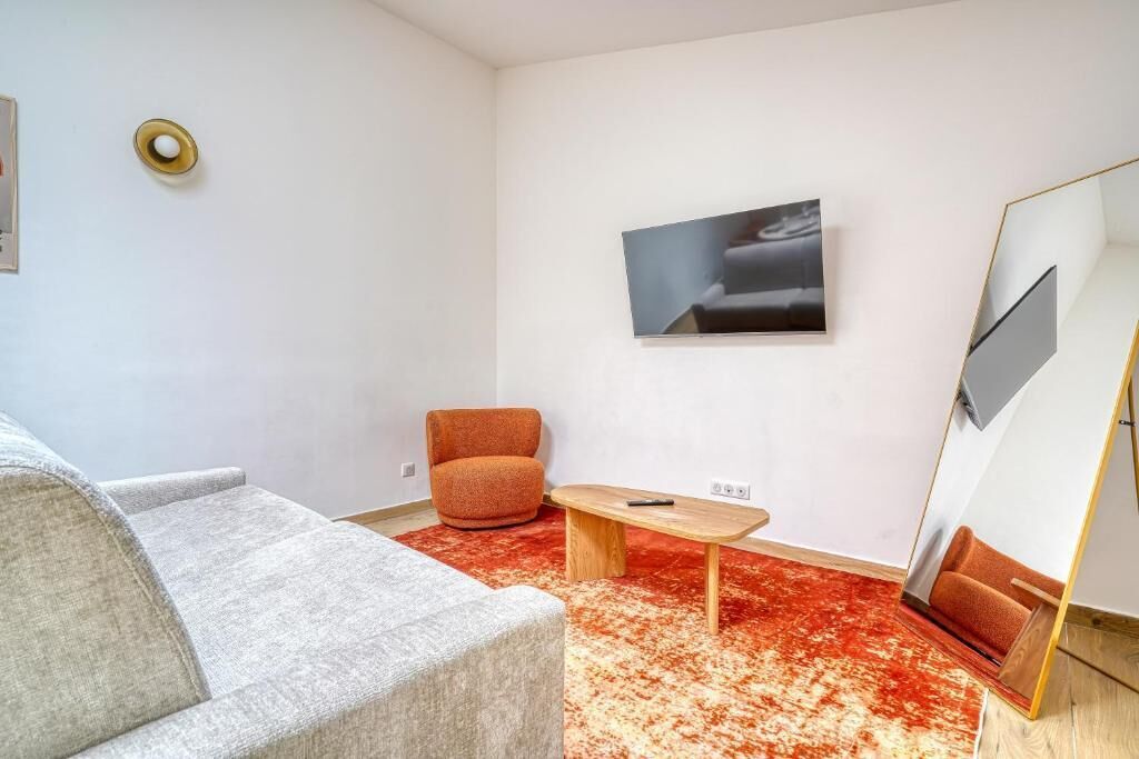 Una sala de estar moderna con un sofá gris, un sillón naranja, una mesa de centro de madera, un tapete rojo con estampado, un televisor de pantalla plana montado en la pared y un espejo grande con marco dorado.