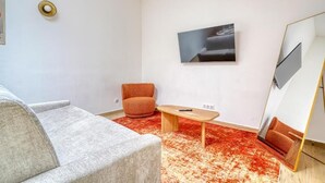 A modern living room with a grey sofa, an orange armchair, a wooden coffee table, a red patterned rug, a flat-screen TV mounted on the wall, and a large mirror with a gold frame.