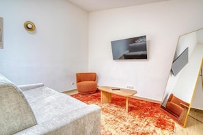 A modern living room with a grey sofa, an orange armchair, a wooden coffee table, a red patterned rug, a flat-screen TV mounted on the wall, and a large mirror with a gold frame.