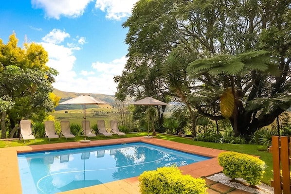 A swimming pool with lounge chairs and umbrellas, surrounded by greenery and hills in the background.