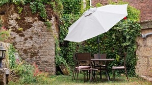 Un espacio al aire libre con mesa y sillas, una sombrilla blanca y una pared de piedra cubierta de vegetación.