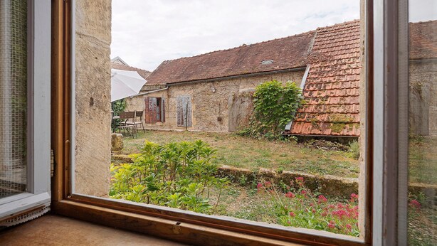 Vista a través de una ventana de un edificio rústico con tejado de baldosas, pared de piedra y un pequeño jardín con diversas plantas y flores.