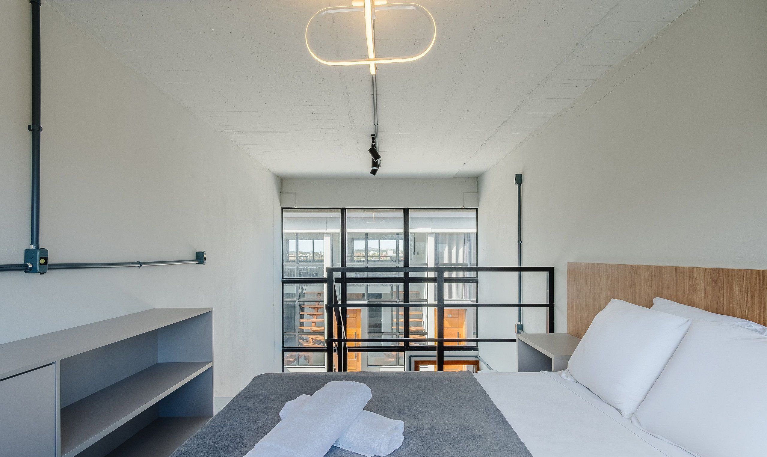 Habitació d'hotel moderna amb un llit, una taula de nit, una estant integrada, vistes a través d'una finestra gran i una lluminària de sostre singular.