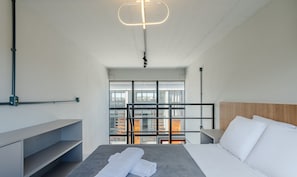 Quarto de hotel moderno com cama, mesa de cabeceira, prateleira embutida, vista através de uma grande janela e luminária de teto original.