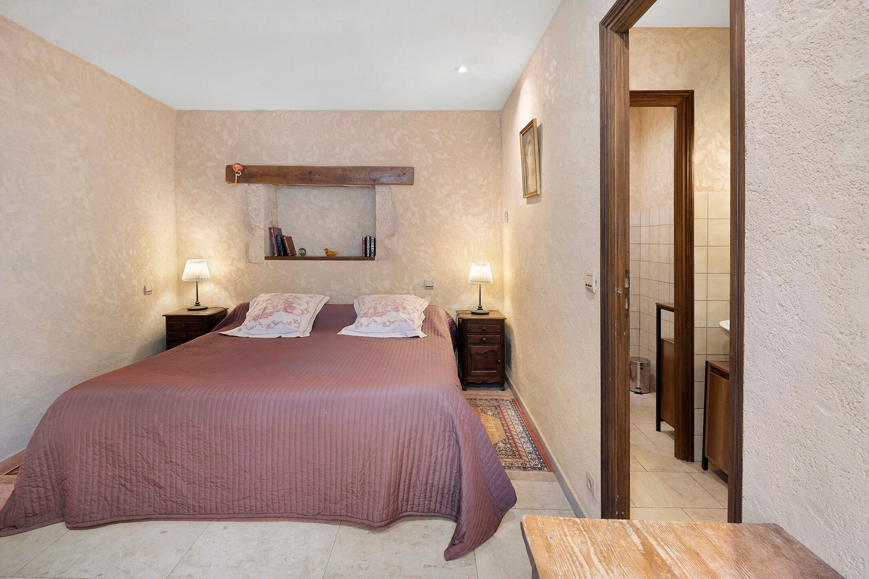 A bedroom with a large bed, two bedside tables, a nightstand, a wall-mounted shelf, and a door leading to another room.