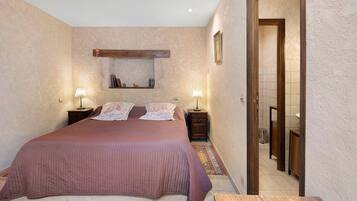 A bedroom with a large bed, two bedside tables, a nightstand, a wall-mounted shelf, and a door leading to another room.