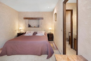 Une chambre avec un grand lit, deux tables de chevet, une commode, une étagère fixée au mur et une porte donnant sur une autre pièce.
