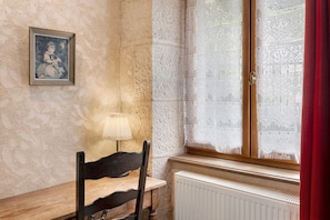 Une pièce comprenant une chaise en bois, une table, un radiateur, un rideau en dentelle à la fenêtre et un tableau encadré accroché au mur.