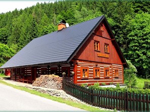 A wooden log cabin with a steeply pitched roof, a chimney, and a stack of firewood in front.