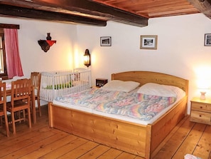 A wooden bed with a colorful quilt, a wooden crib, a wooden nightstand, and a wall-mounted lamp.