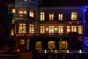 Un edificio de varios pisos con ventanas amplias y balcones, un espacio de estar al aire libre techado con mesas y sillas, y un puente que conecta el edificio con la propiedad contigua.