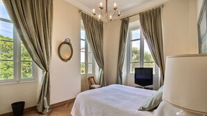 A bedroom with a bed, a television, a chair, a mirror, and a chandelier. - Classic South of France elegances set in an oasis in the heart of Cannes (Cannes)