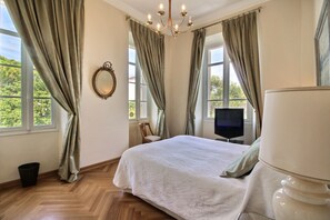 A bedroom with a bed, a television, a chair, a mirror, and a chandelier. - Classic South of France elegances set in an oasis in the heart of Cannes (Cannes)