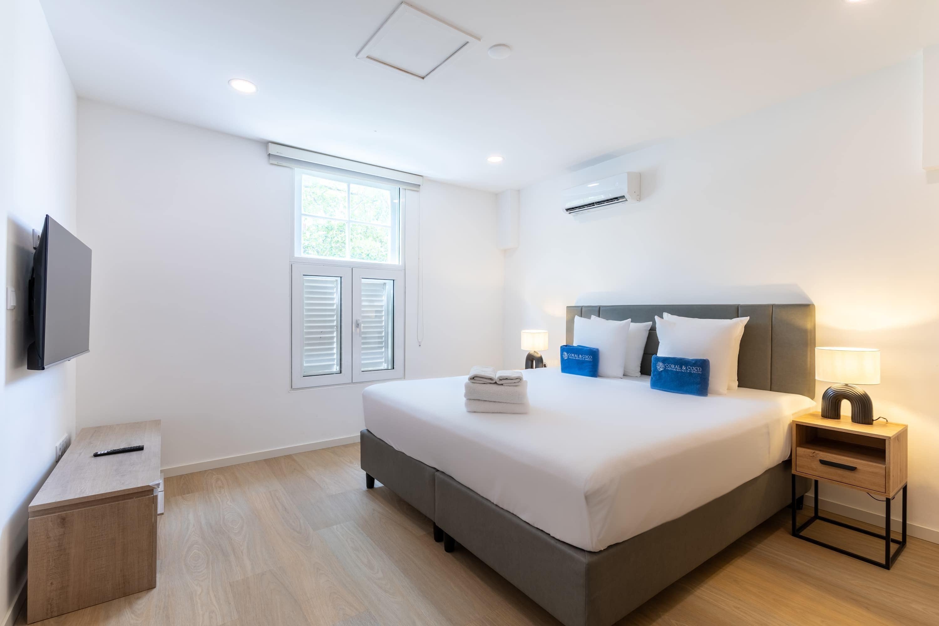 Habitación de hotel con cama, televisor, ventana con persianas, mesita de noche con lámpara y un pequeño armario de madera.
