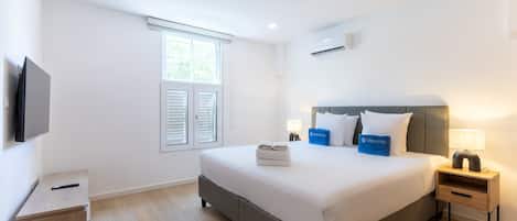 Une chambre d’hôtel comprenant un lit, une télévision, une fenêtre avec des stores, une table de chevet avec une lampe et une petite armoire en bois.