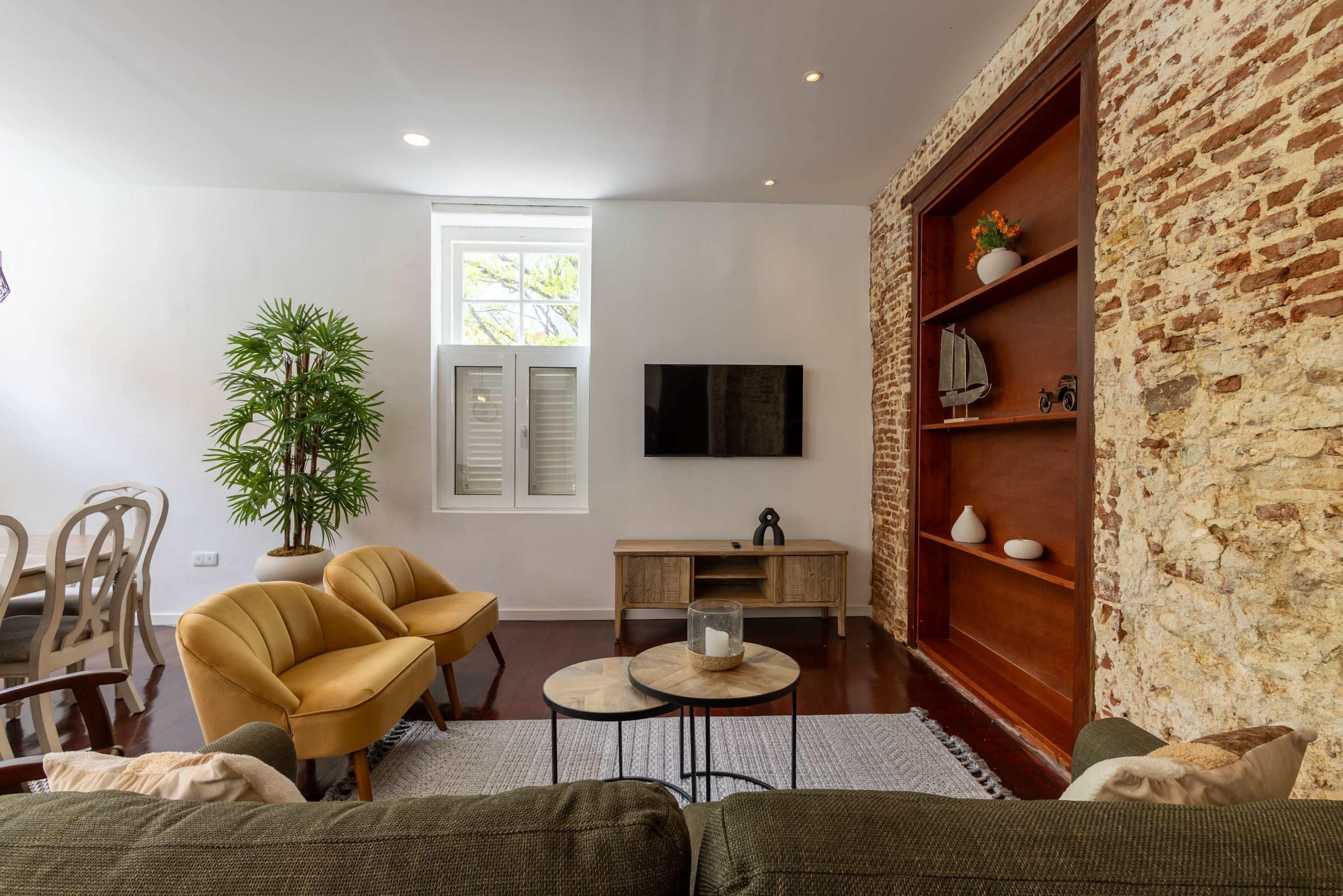 Ein Wohnzimmer mit Ziegelwand, Fernseher, Pflanze, zwei gelben Sessel, Couchtisch aus Holz und einer Couch.