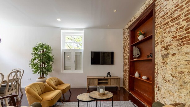 A living room with a brick wall, a television, a plant, two yellow chairs, a wooden coffee table, and a sofa. - NEW Beautifully Restored Historic Apartment (Willemstad)