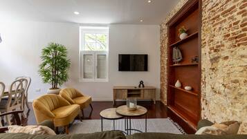 Un salón con pared de ladrillo, televisor, planta, dos sillas amarillas, una mesa de centro de madera y un sofá.