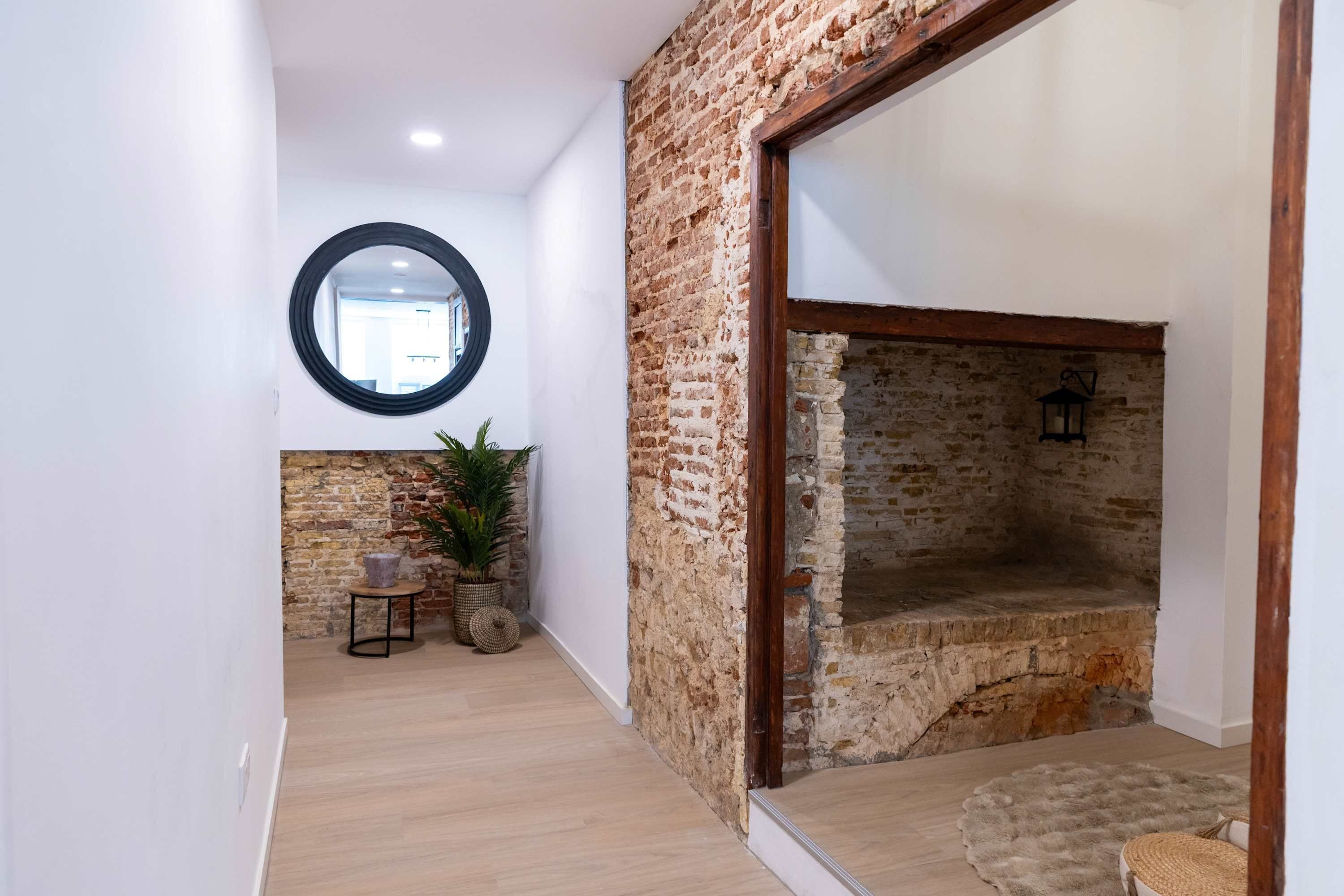 Un pasillo con piso de madera, pared de ladrillo, un espejo redondo y un banco de piedra.