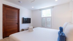 A bedroom with a wooden door, a flat-screen TV, a window with shutters, a bed with white linens, and a blue towel. - NEW Beautifully Restored Historic Apartment (Willemstad)