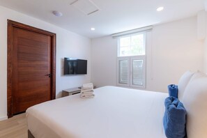 A bedroom with a wooden door, a flat-screen TV, a window with shutters, a bed with white linens, and a blue towel. - NEW Beautifully Restored Historic Apartment (Willemstad)
