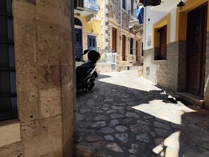 Eine schmale Kopfsteinpflasterstraße mit einem abgestellten Roller am Rand, Gebäude mit Steinfassaden und einem klaren Himmel.