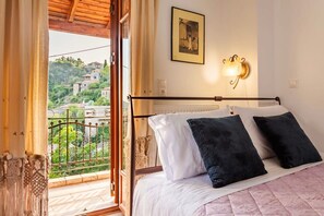 A bedroom with a bed, a balcony with a view of buildings, and a wall-mounted light fixture. - Stemnitsa Stone Residence - Cosy Mountain Escape (Gortynia)