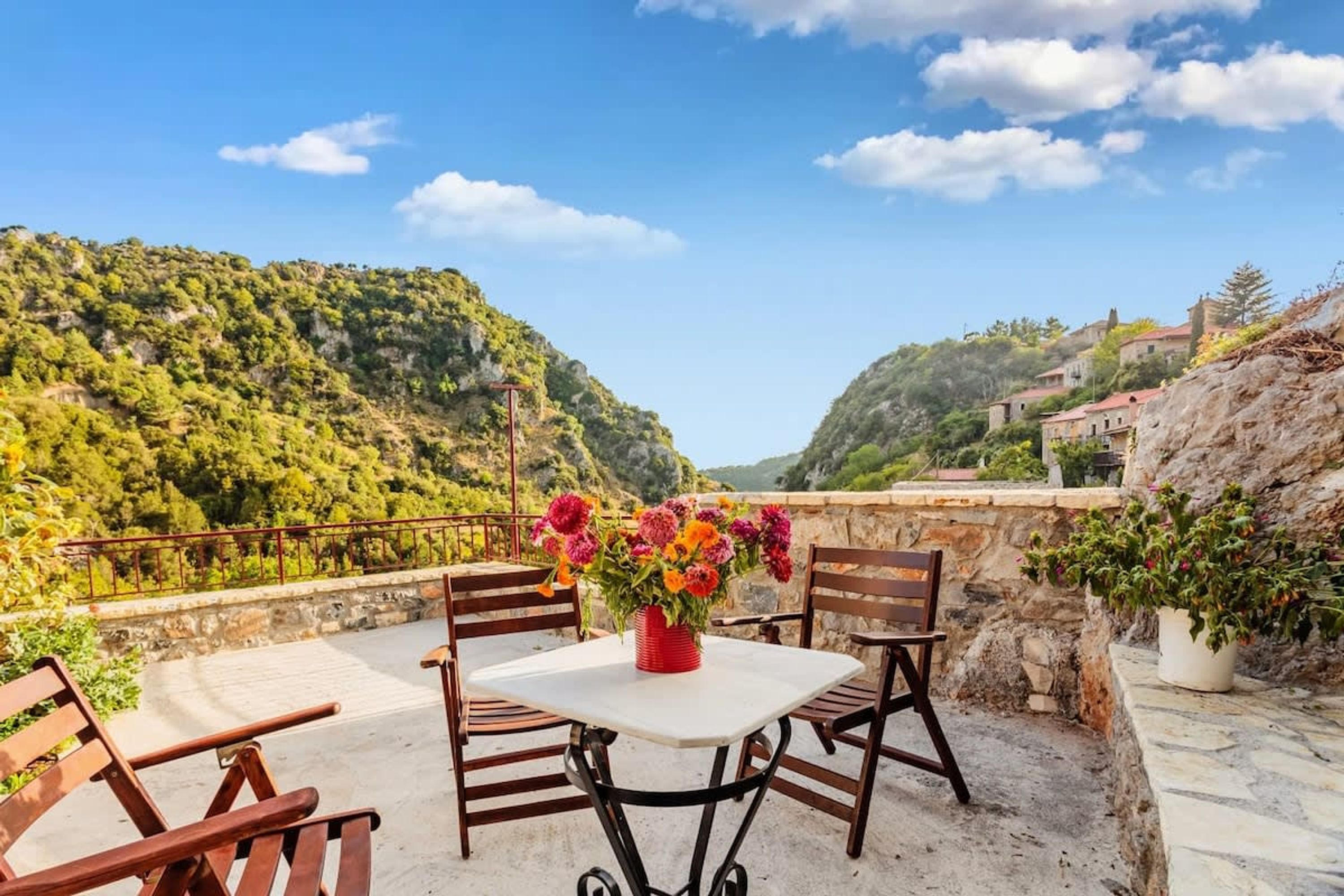 An outdoor seating area with a table and chairs, a vase of flowers, and a view of a hillside.