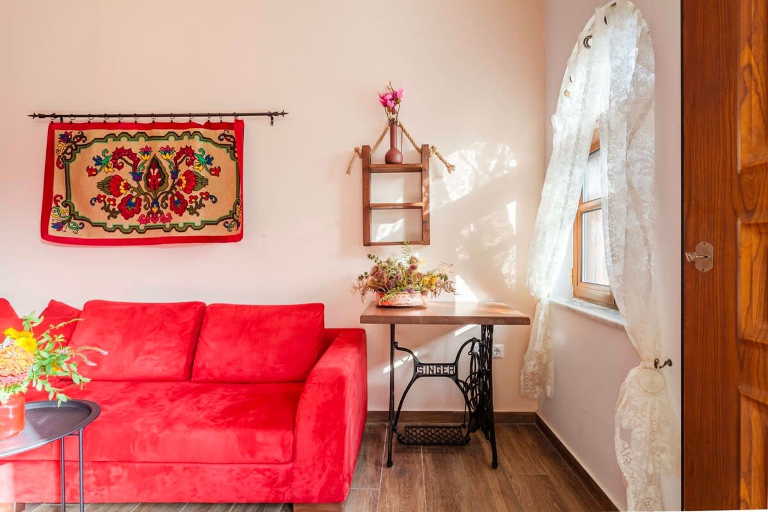 A room with a red sofa, a wooden table with a vase of flowers, a sewing machine, and a window with a white curtain.