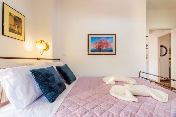 A hotel room with a bed, pillows, a quilted pink bedspread, a framed painting, and a wall-mounted light fixture. - Stemnitsa Stone Residence - Cosy Mountain Escape (Gortynia)