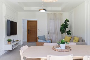 Ein modernes Wohnzimmer mit Flachbildfernseher, einem Esstisch mit Stühlen, einer Couch mit Kissen und einer Zimmerpflanze.