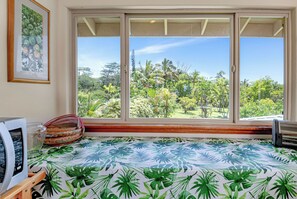 Ein Zimmer mit großem Fenster, einem Bett mit tropischem Bettbezug, einem gerahmten Bild an der Wand und einem kleinen Tisch mit einem gewebten Korb.