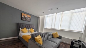 A modern bedroom with a bed, a gray sofa, and a large window with blinds. - City View Studio Apartment – Liverpool (Liverpool)
