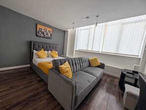 A modern bedroom with a bed, a gray sofa, and a large window with blinds. - City View Studio Apartment – Liverpool (Liverpool)