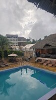 Una piscina con sillas de descanso y sombrillas, un área de estar techada y un edificio con techo de paja al fondo.