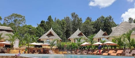 A pool area with thatched-roof bungalows, lounge chairs, and umbrellas.