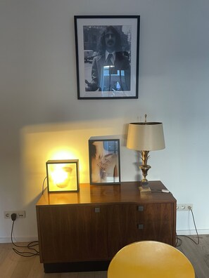 Ein Holzkabinent mit einem gerahmten Bild, einer beleuchteten Bilderleiste und einer Tischlampe vor einer schlichten Wand.