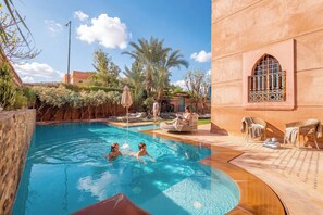 A swimming pool with two people in the water, surrounded by palm trees and a building with a distinctive window. - Villa Taj Sofia & Spa (Marrakech)